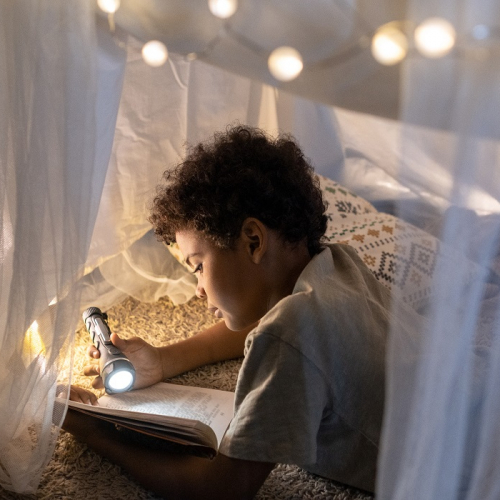 Les Nuits de la lecture 2025 dans le Rhône : Un petit garçon lit un livre à la lampe torche sous une tente