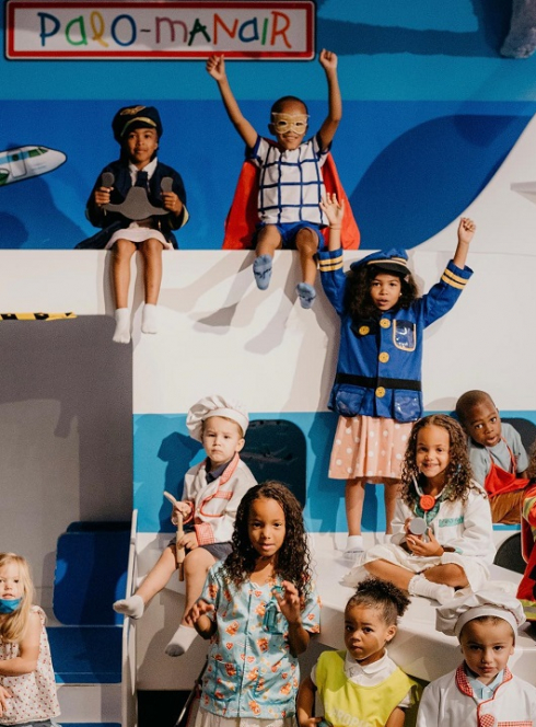 Palomano : Des enfants portent les tenues de diverses professions dans une salle qui ressemble à un aéroport