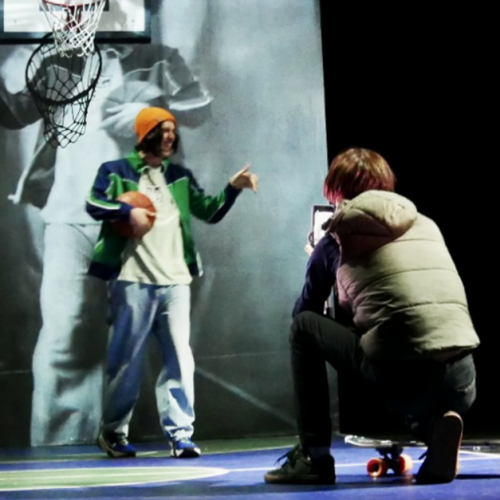 To like or not - Cie Ex Voto à la lune : Un ado sur un banc avec son téléphone et un autre qui prend en photo un jeune avec un ballon de basket