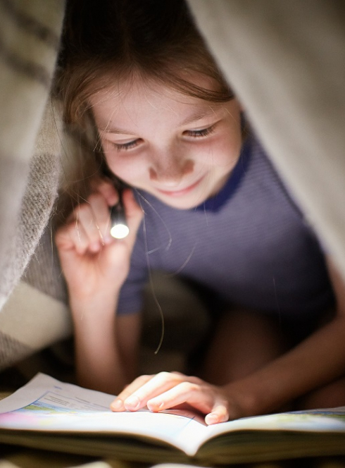Les Nuits de la lecture : Une petite fille lit un livre sous un plaid avec une lampe torche