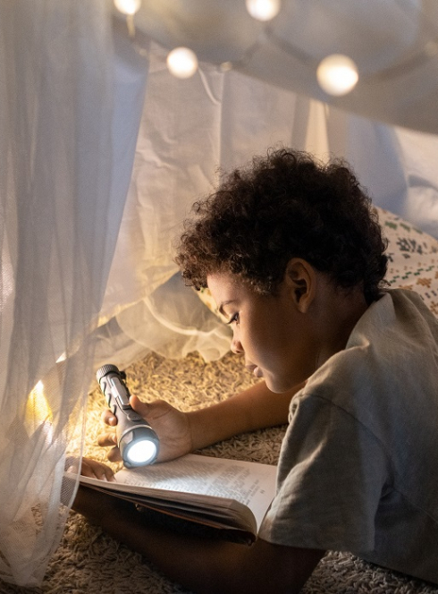 Les Nuits de la lecture 2025 dans le Rhône : Un petit garçon lit un livre à la lampe torche sous une tente